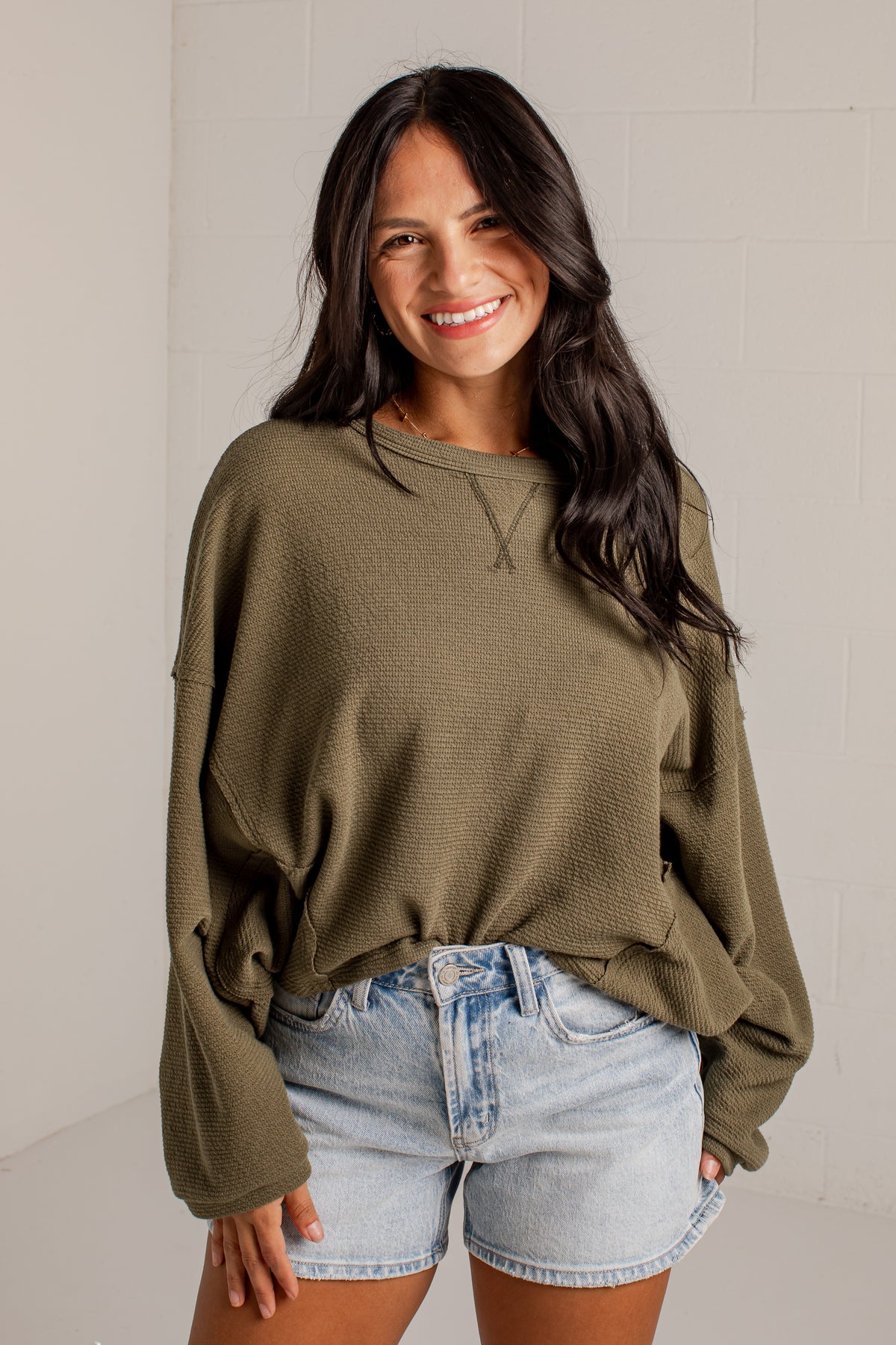 A woman wearing a textured olive green long-sleeve crop top and light-wash denim shorts, standing against a light gray brick wall.