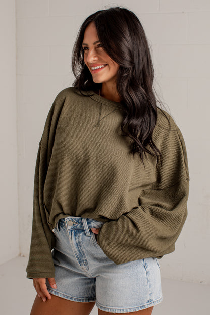 A woman wearing an oversized olive green waffle-knit long-sleeve sweatshirt and light-wash denim shorts, standing against a white brick wall.