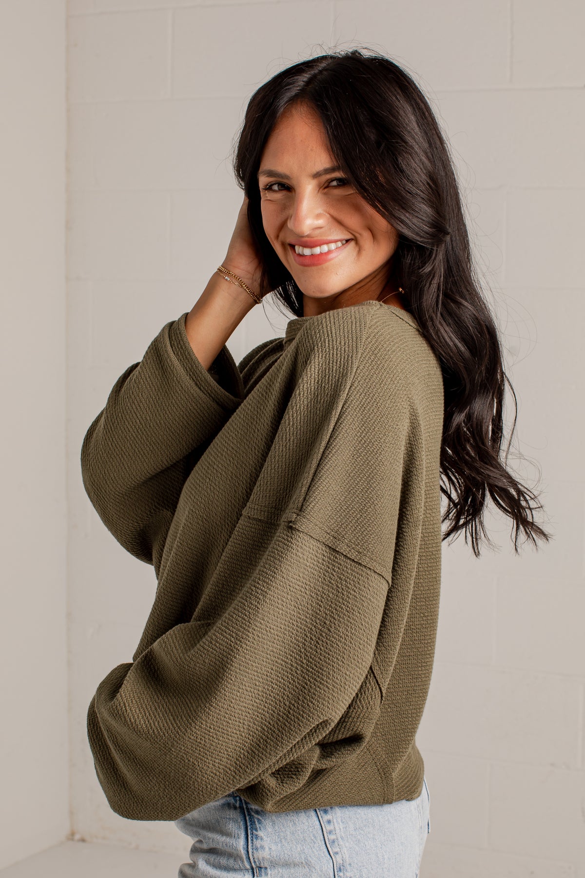 A woman with long dark hair smiling and looking over her shoulder, wearing an oversized olive green waffle-knit sweatshirt and light blue jeans, standing against a light gray brick wall.