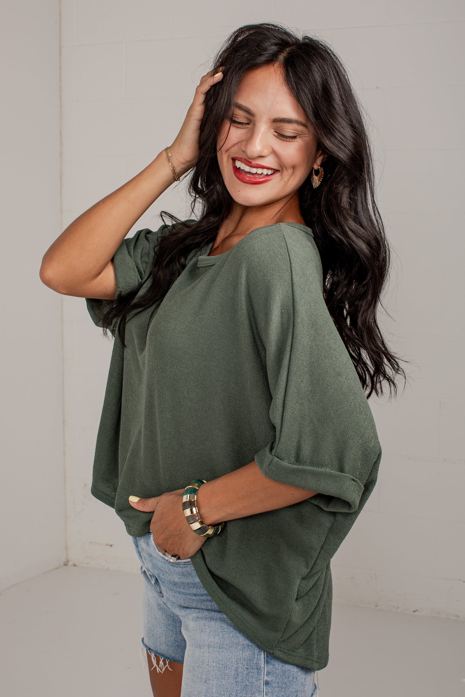 A woman wearing a loose, short-sleeve olive green top and light blue denim shorts, posing in a bright studio setting.
