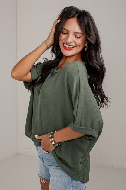 A woman wearing a loose, short-sleeve olive green top and light blue denim shorts, posing in a bright studio setting.