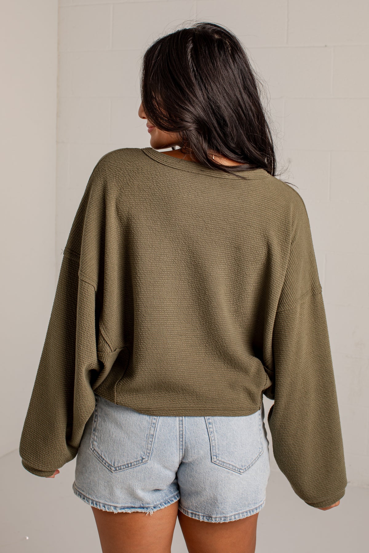 A woman shown from behind wearing a textured olive green cropped sweater and light-wash denim shorts against a neutral off-white wall.