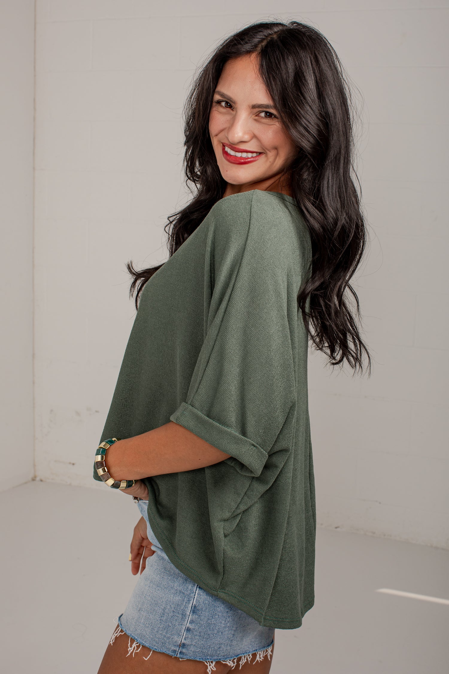A woman with long dark hair is shown from the back and side, wearing an olive green oversized short-sleeve top with rolled cuffs and light blue denim shorts in a bright indoor setting.