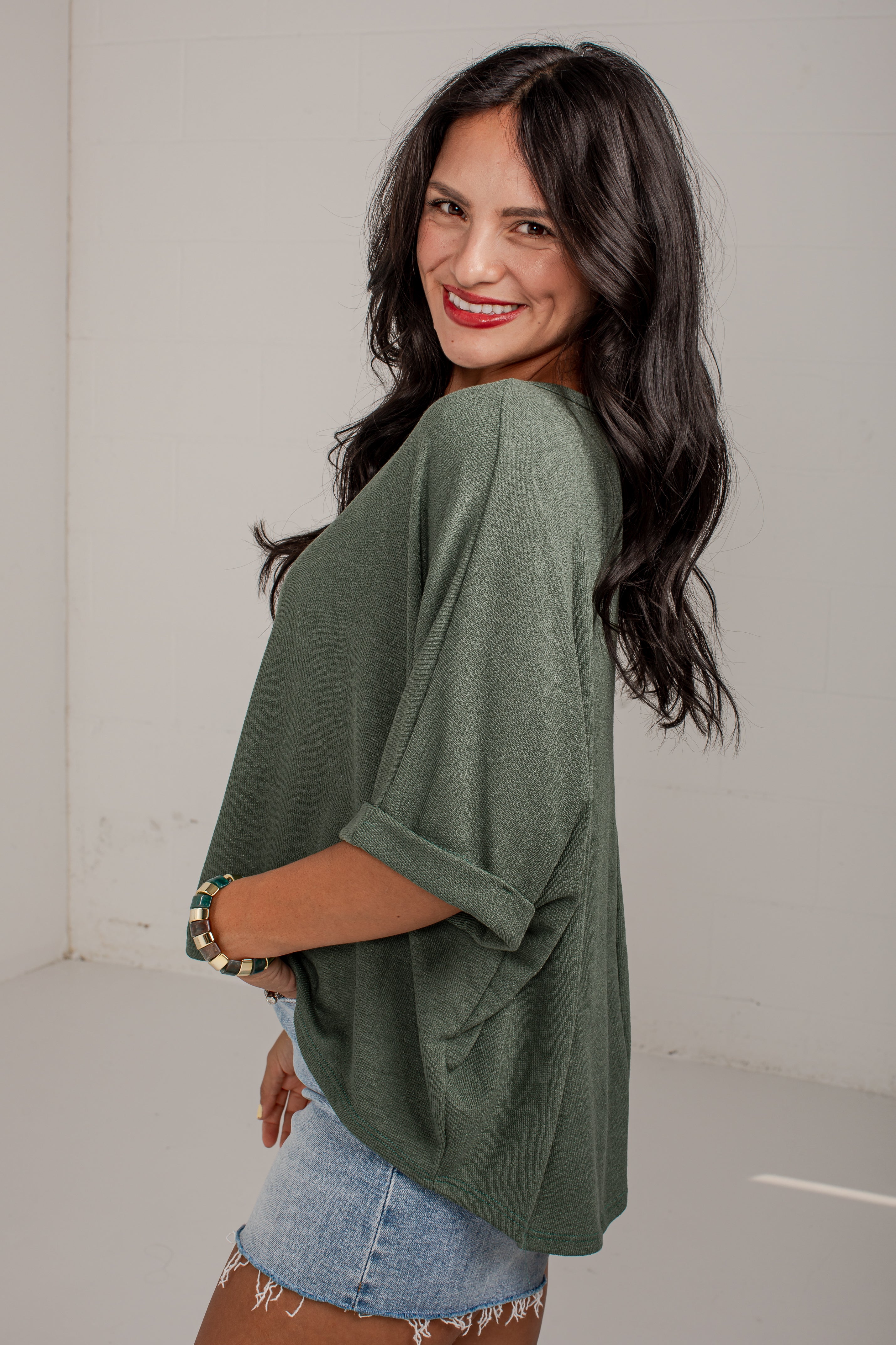 A woman with long dark hair is shown from the back and side, wearing an olive green oversized short-sleeve top with rolled cuffs and light blue denim shorts in a bright indoor setting.