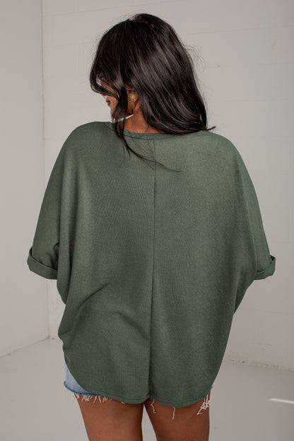 A woman viewed from behind wearing an oversized sage green knit top with rolled sleeves and a split back seam, paired with denim shorts.
