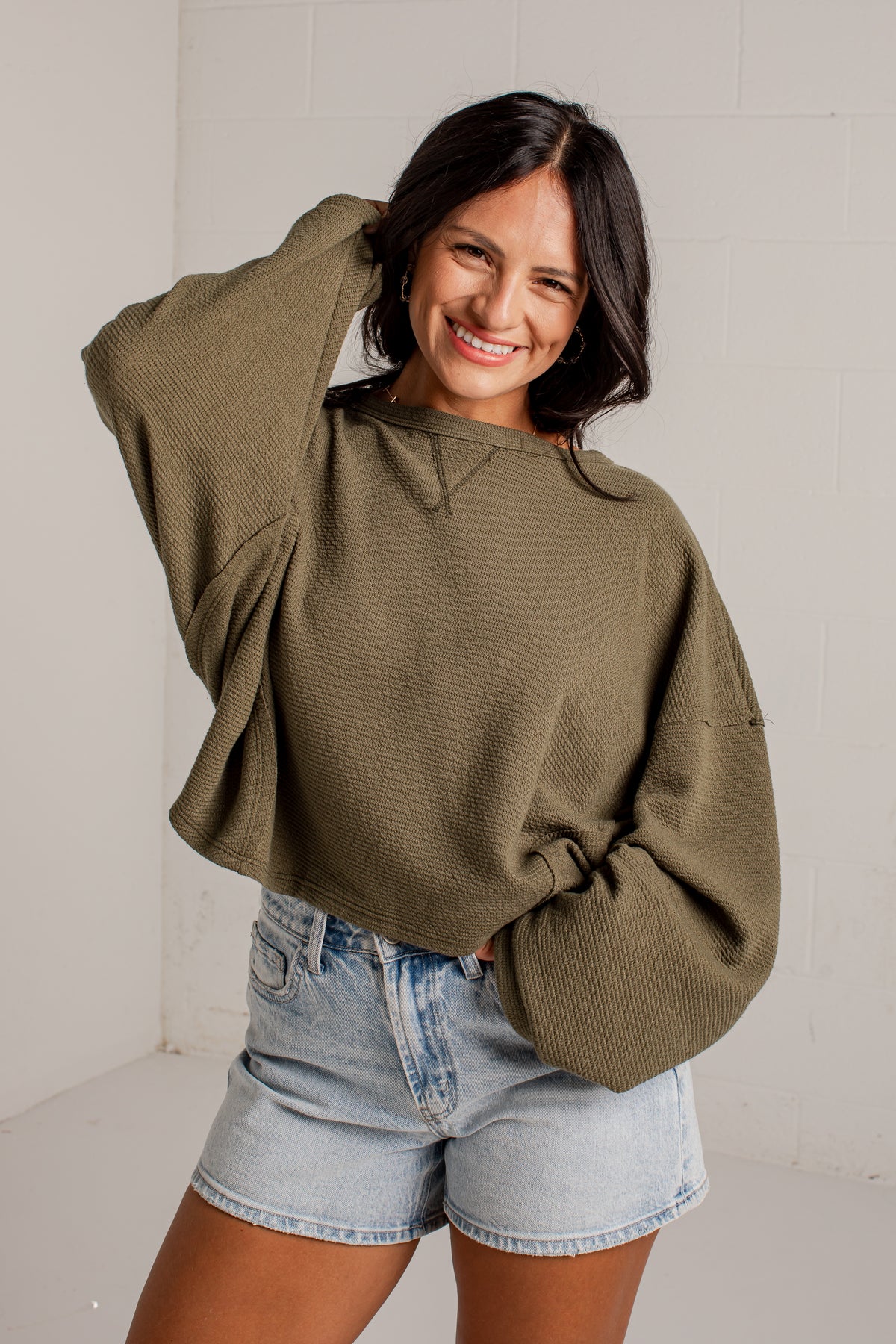 A woman wearing an oversized olive green knit sweater and light-wash denim shorts, posing in front of a white brick wall.