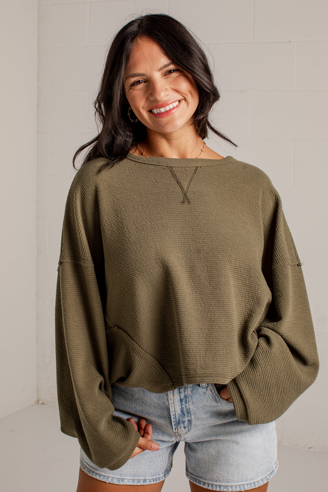 A woman smiling while wearing a textured olive green long-sleeve crop top sweater and light-wash denim shorts against a white brick wall background.
