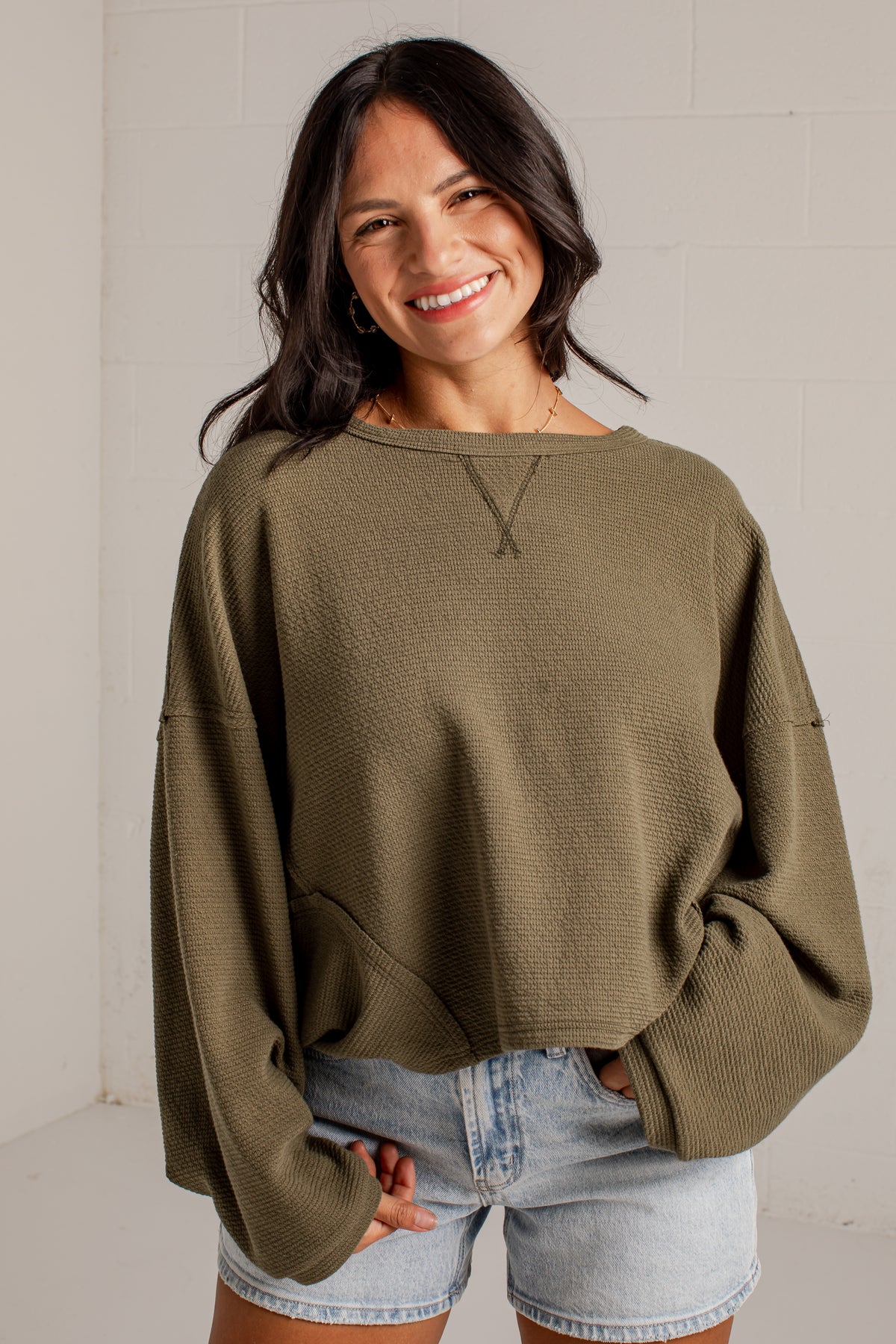 A woman smiling while wearing a textured olive green long-sleeve crop top sweater and light-wash denim shorts against a white brick wall background.