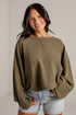 A woman smiling while wearing a textured olive green long-sleeve crop top sweater and light-wash denim shorts against a white brick wall background.