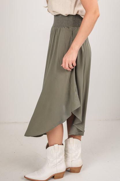 Person wearing a long olive green skirt with white boots on a plain background