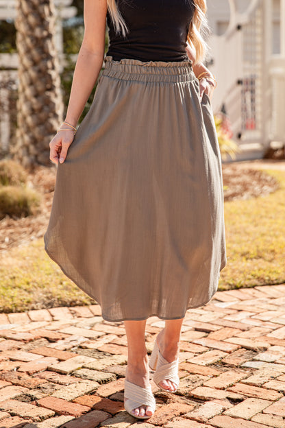 Person wearing a beige skirt and black top on a brick path with grass and trees in the background