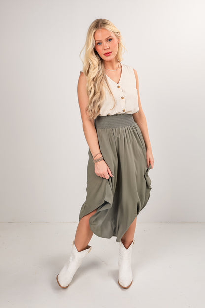 Woman wearing a white sleeveless top and green skirt with white boots on a plain background