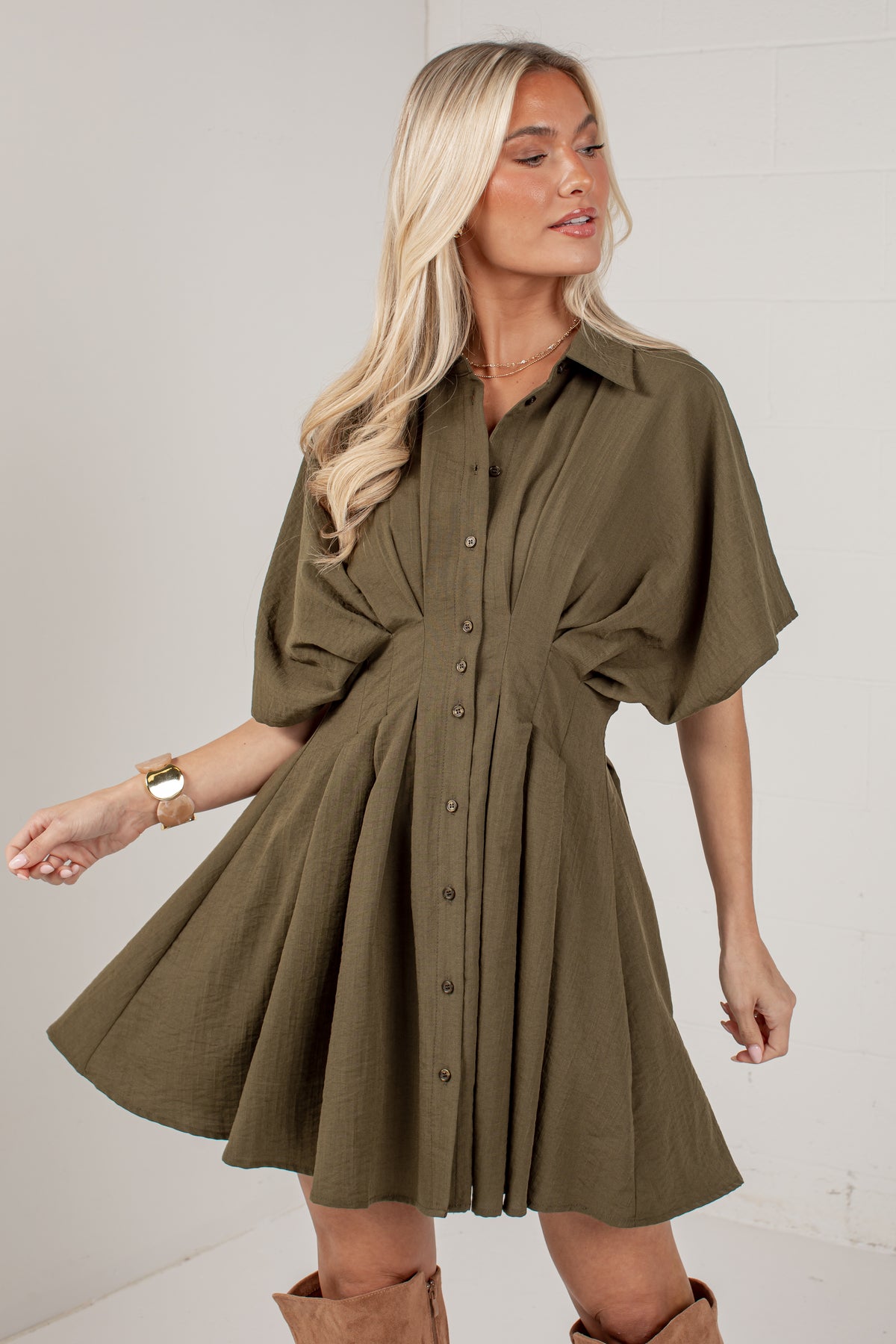 A woman modeling an olive green short-sleeve button-up dress with a flared skirt, shown against a light gray wall.