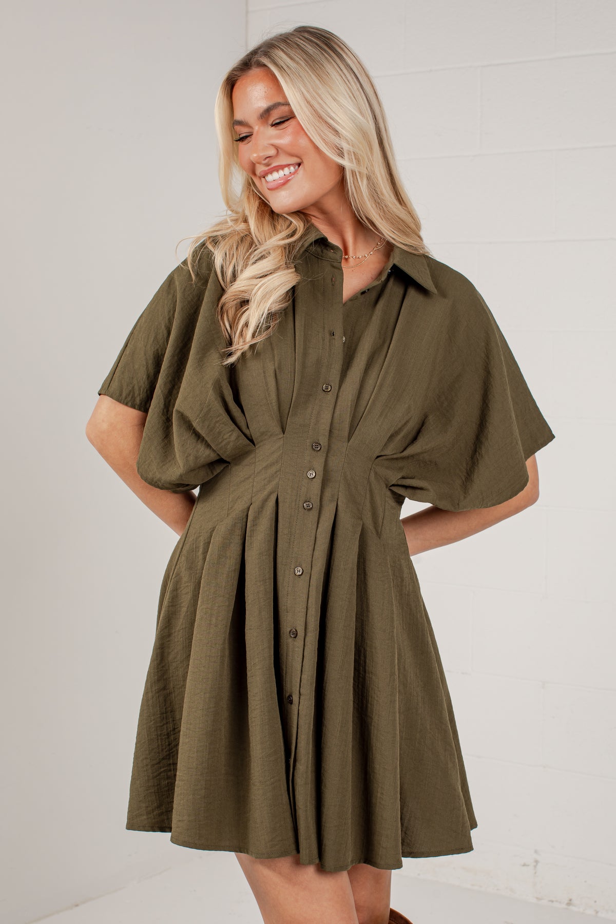 A woman wearing an olive green short-sleeve button-up dress with a gathered waist and flared skirt, standing against a white paneled wall.