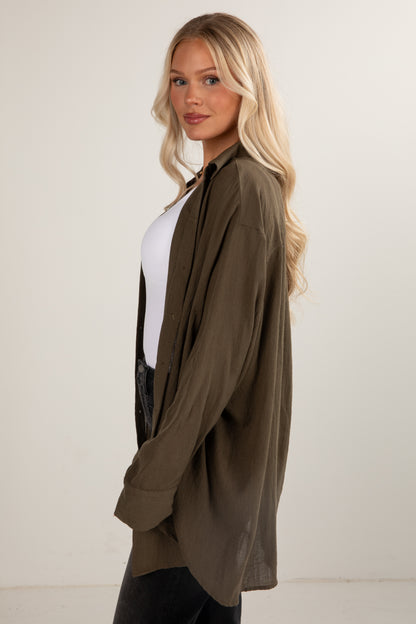 Woman wearing an olive green cardigan over a white shirt and black pants on a plain background