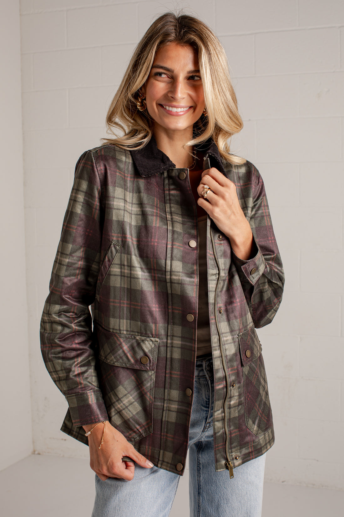 A woman wearing a dark olive green and burgundy plaid button-up jacket with a dark collar, paired with light blue jeans, standing against a white brick wall.