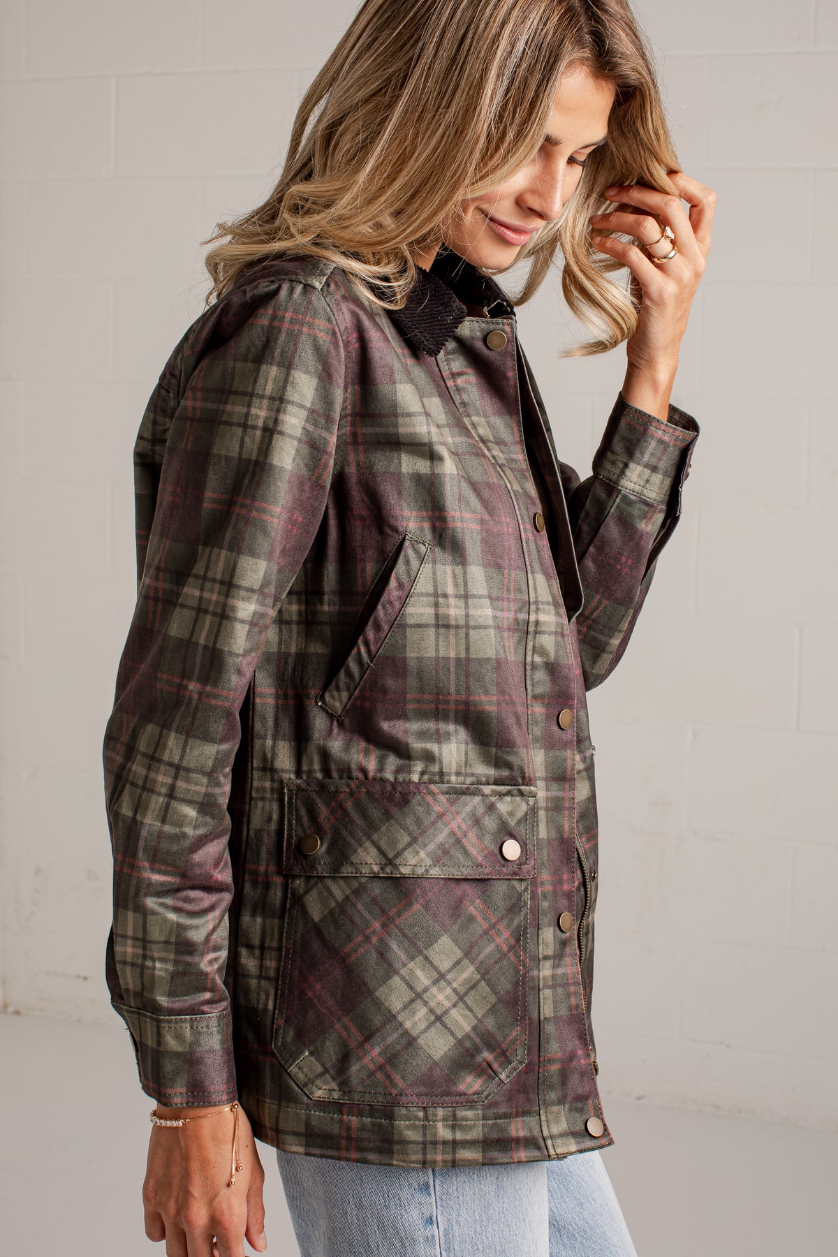 A woman wearing a long-sleeve plaid waxed jacket in muted green, brown, and burgundy tones, paired with light blue jeans, photographed against a light gray brick wall.