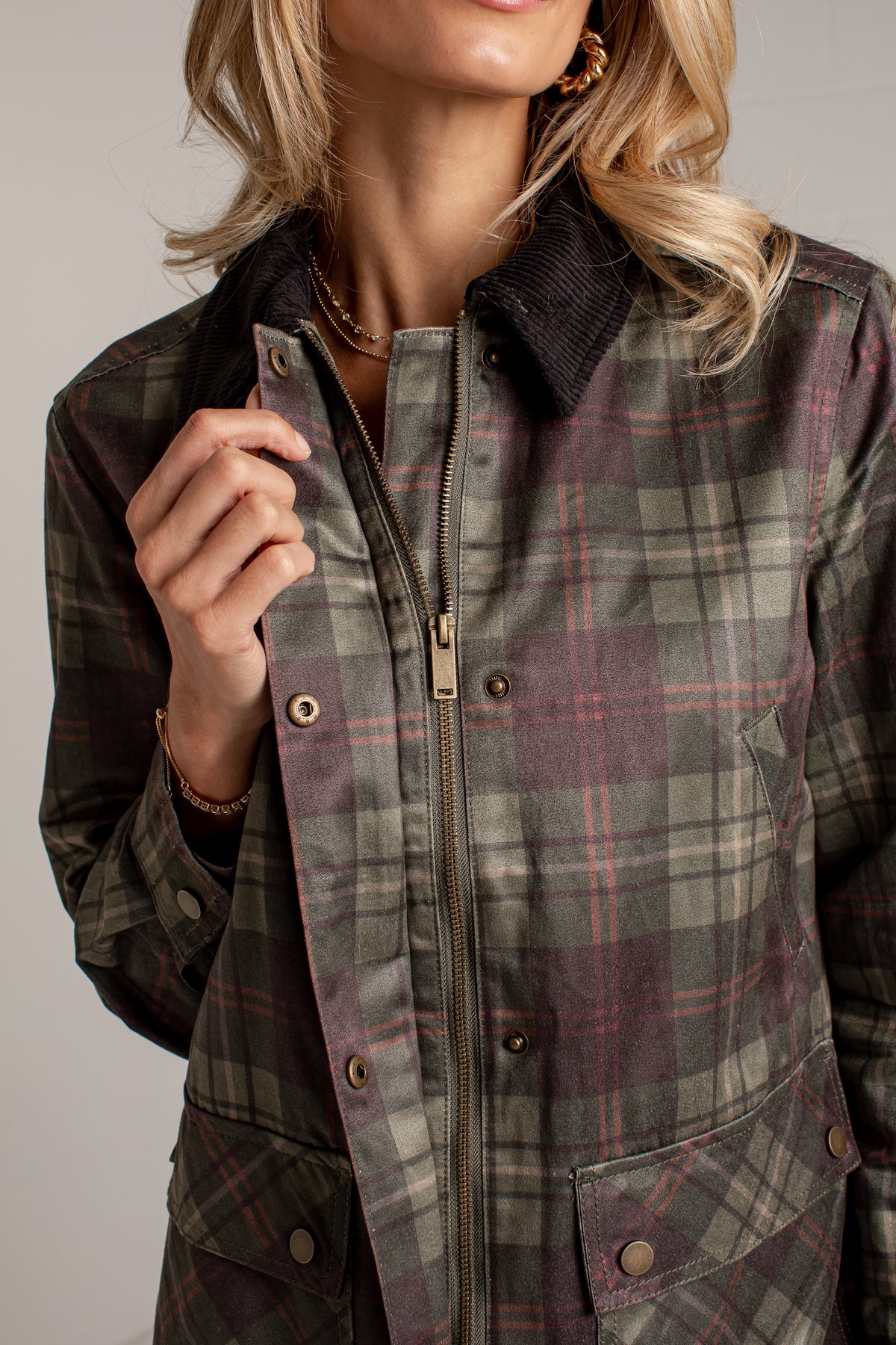 A close-up of a woman wearing a plaid zip-front jacket with a corduroy collar. The jacket features a muted plaid pattern in olive green, gray, and burgundy with brass-tone snaps and a zipper.