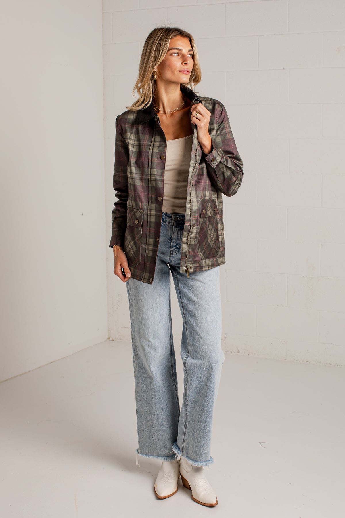 A woman modeling a dark green and brown plaid button-up shirt jacket over a beige top, paired with light-wash wide-leg jeans and white ankle boots, standing in a bright white studio corner.