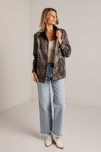 A woman modeling a dark green and brown plaid button-up shirt jacket over a beige top, paired with light-wash wide-leg jeans and white ankle boots, standing in a bright white studio corner.