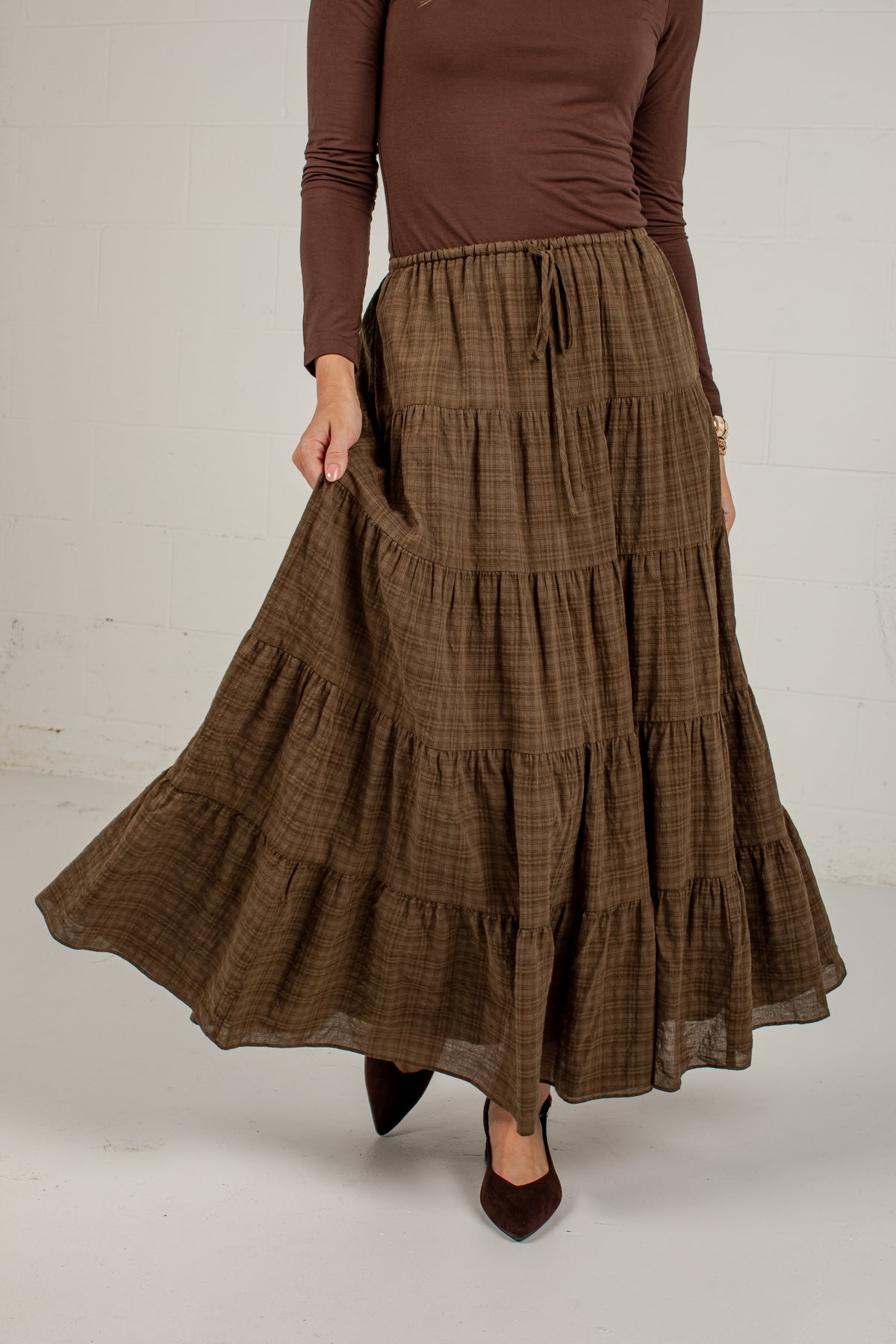A model wearing a brown tiered maxi skirt with a subtle plaid pattern, paired with a matching brown long-sleeve top and brown pointed-toe heels, standing against a white wall.