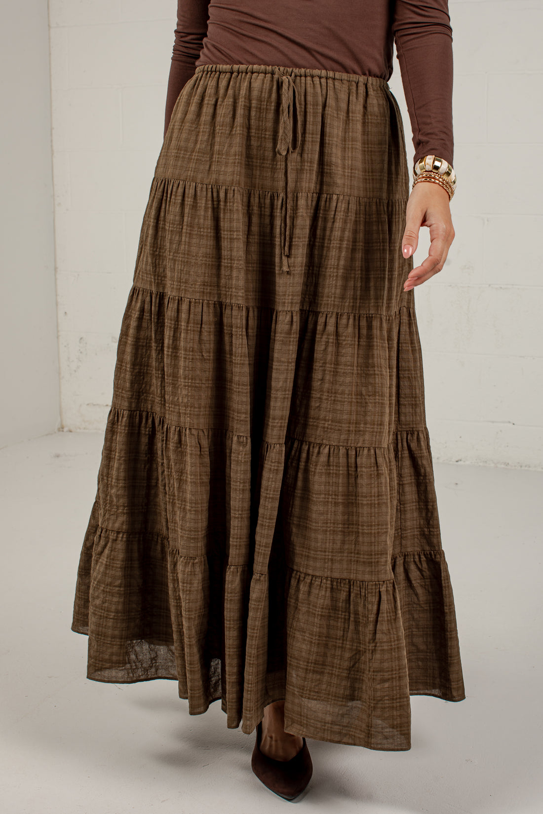 A brown tiered maxi skirt with a subtle plaid pattern, worn by a model with a brown long-sleeve top and dark brown pointed-toe heels.
