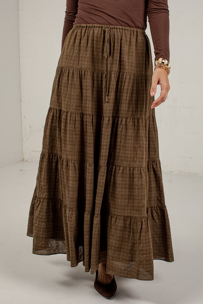 A brown tiered maxi skirt with a subtle plaid pattern, worn by a model with a brown long-sleeve top and dark brown pointed-toe heels.