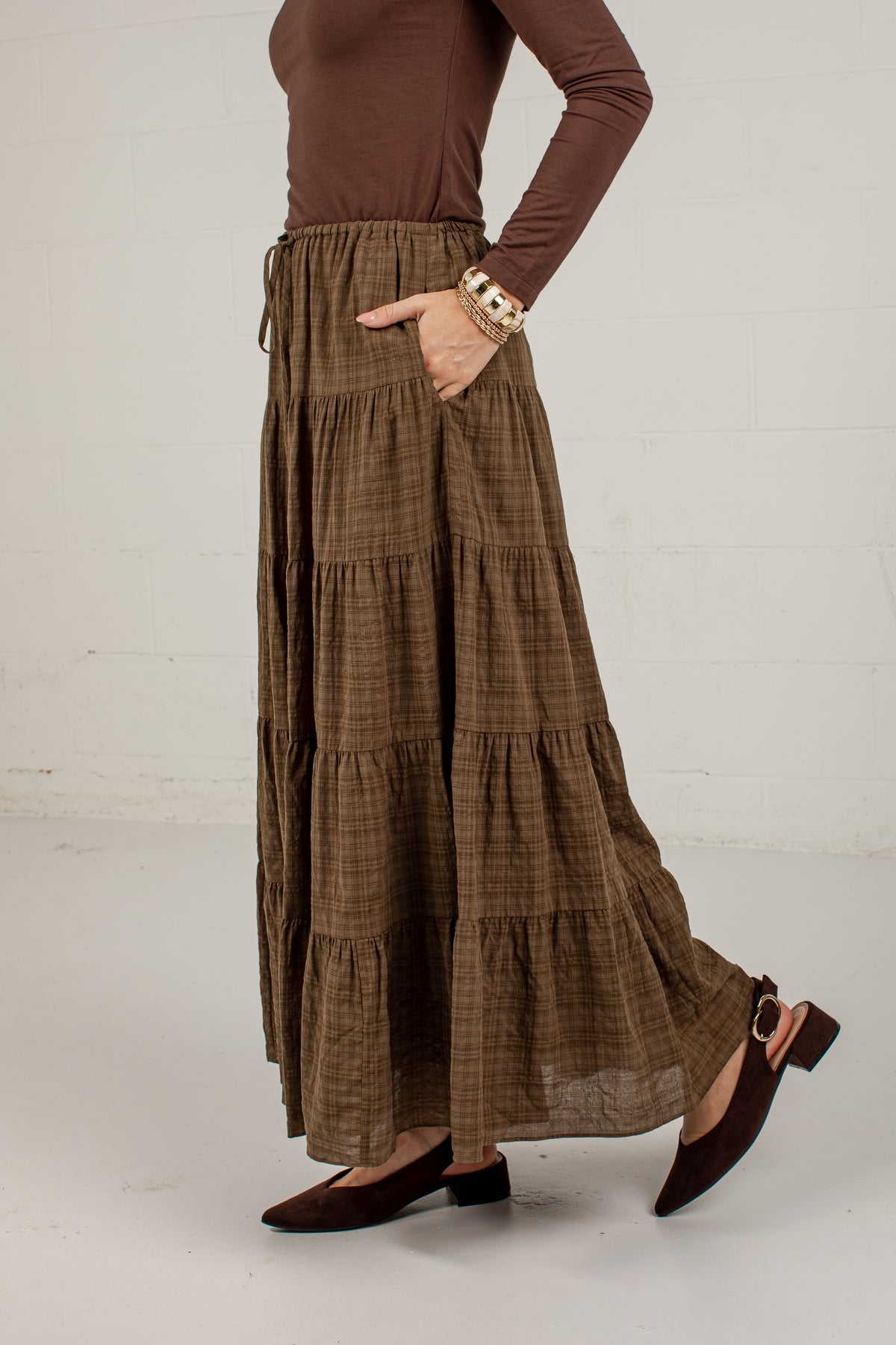 A model wearing a brown and beige plaid tiered maxi skirt with pockets, paired with a brown long-sleeve top and brown flat shoes, standing against a white brick wall.