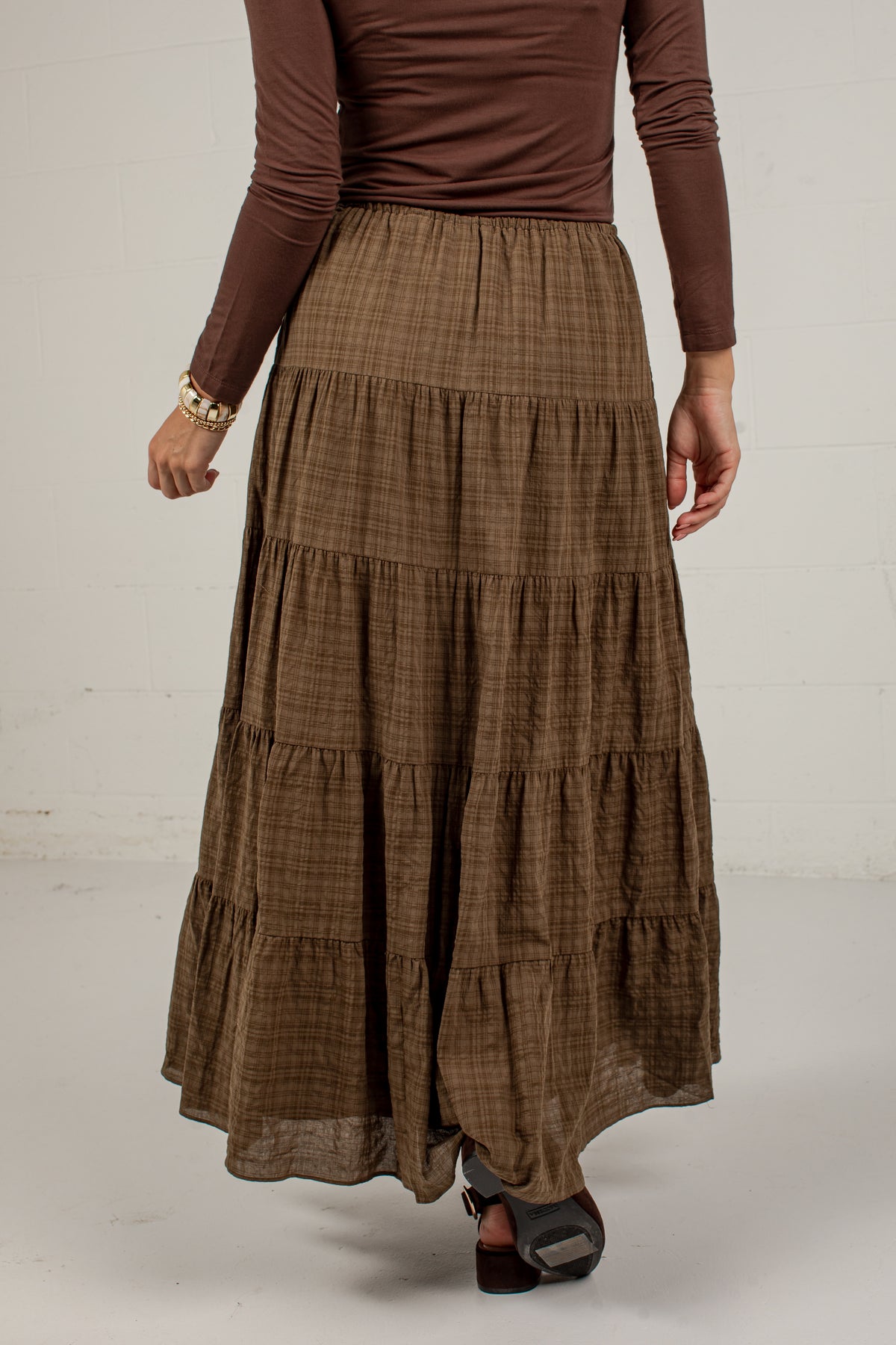 A model wearing a brown tiered maxi skirt with a subtle plaid pattern, paired with a matching brown long-sleeve top and dark sandals, shown from the waist down against a white studio background.