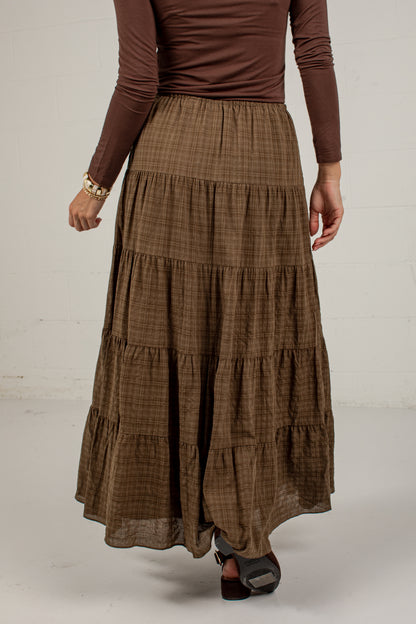 A model wearing a brown tiered maxi skirt with a subtle plaid pattern, paired with a matching brown long-sleeve top and dark sandals, shown from the waist down against a white studio background.