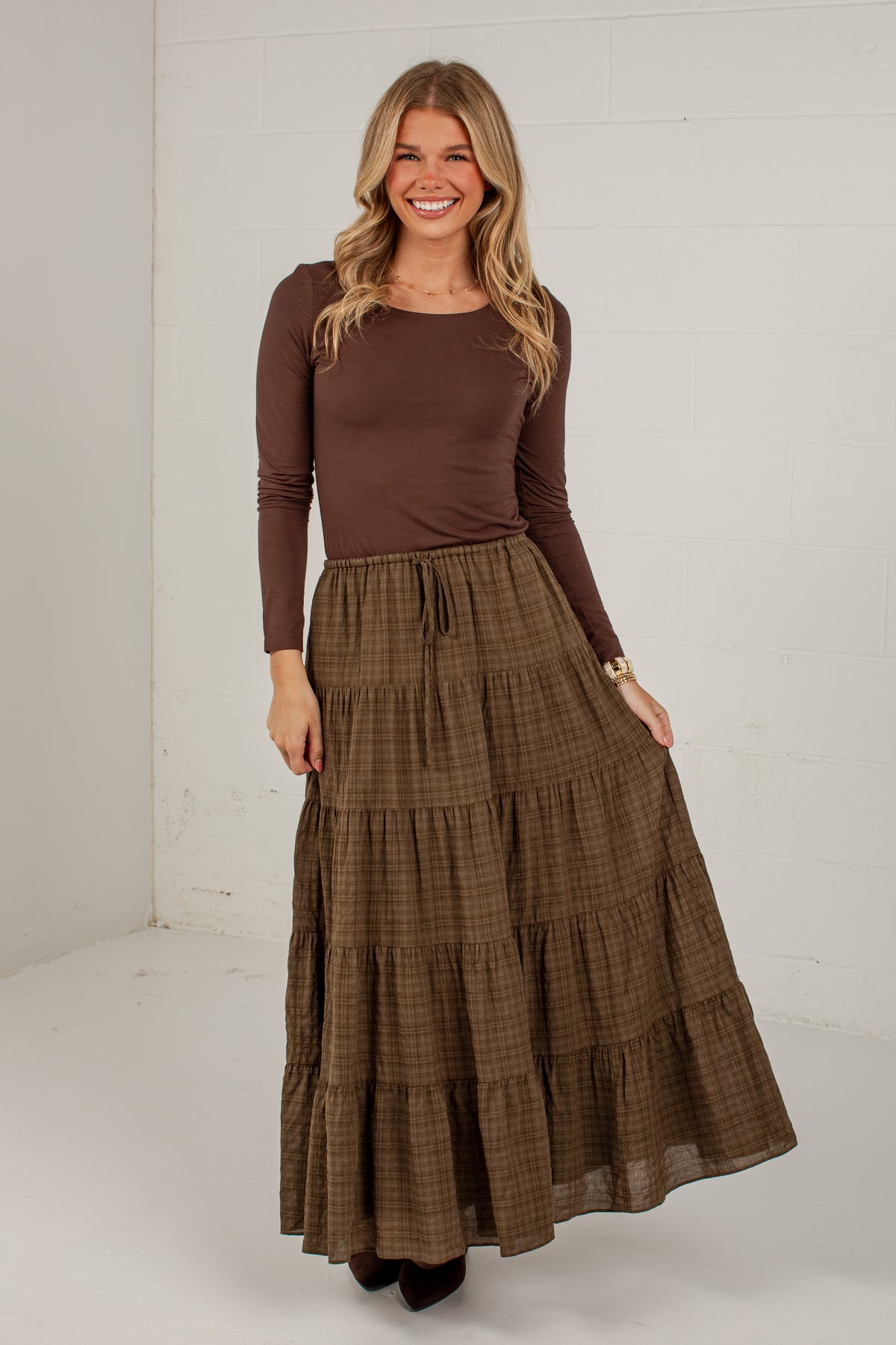 A woman modeling a brown long-sleeve top and a brown tiered plaid maxi skirt, standing against a white studio wall.