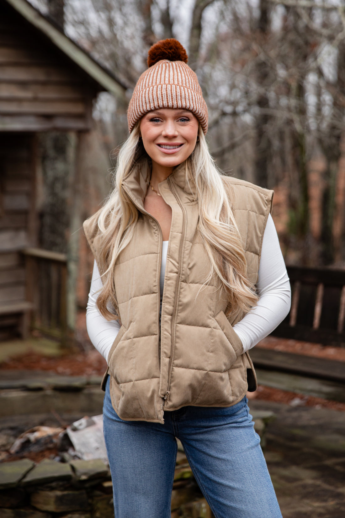 Woman wearing a beige puffer vest and beanie outdoors near a wooden cabin.