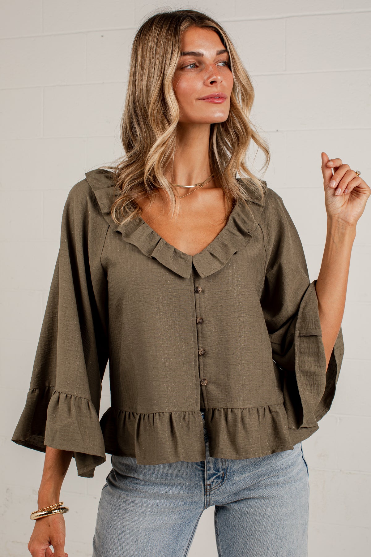 A woman wearing an olive green ruffled blouse with three-quarter bell sleeves and a button-down front, paired with light blue jeans, standing against a white brick wall.