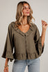 A woman wearing an olive green ruffled blouse with three-quarter bell sleeves and a button-down front, paired with light blue jeans, standing against a white brick wall.