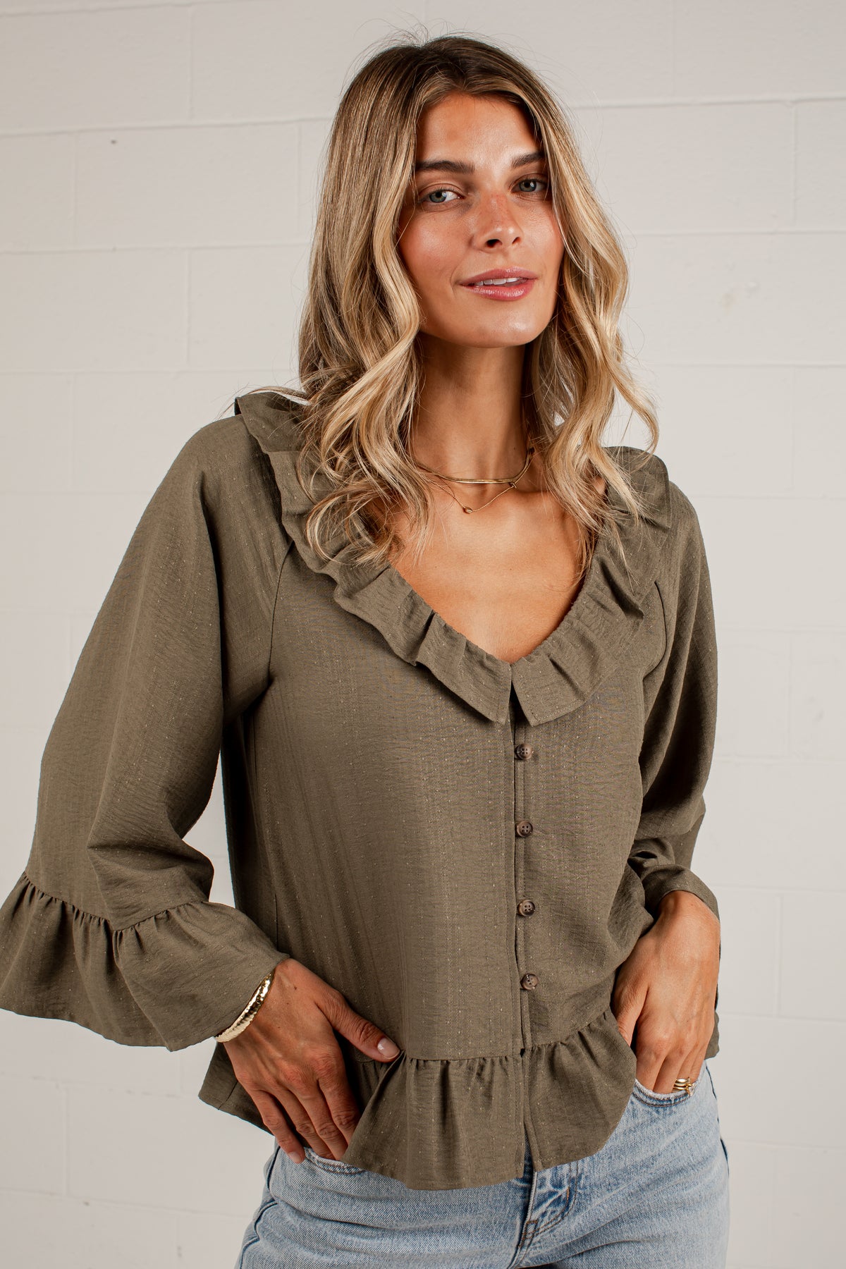 A woman wearing an olive green long-sleeve blouse with ruffle trim, a button-down front, and ruffled cuffs, paired with light-wash blue jeans against a white brick wall.