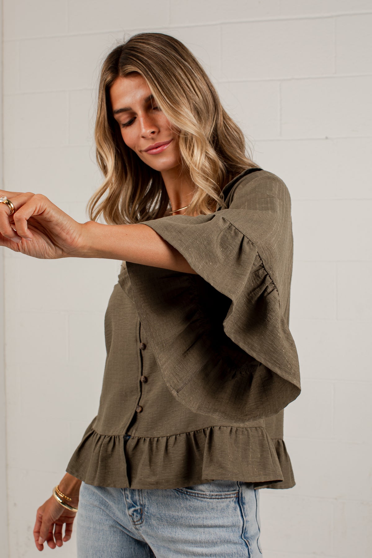 A woman wearing an olive green linen blouse with wide flutter sleeves, a button-down front, and a ruffled hem, paired with light blue jeans against a white brick wall.