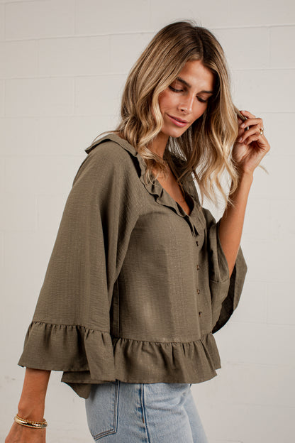 A woman wearing an olive green blouse with ruffled trim and light blue jeans, standing against a white brick wall.