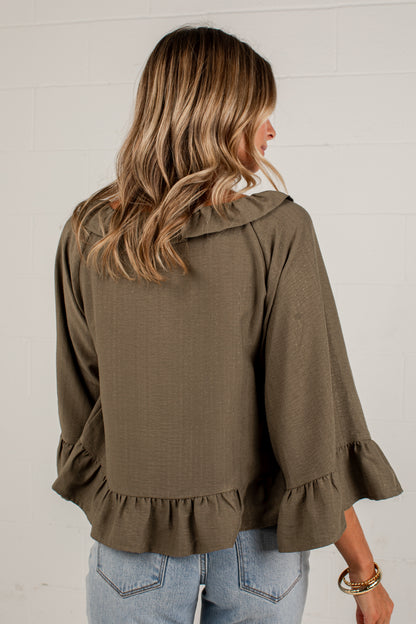 A woman shown from the back wearing an olive brown ruffled blouse with three-quarter bell sleeves and light blue jeans, standing against a white brick wall.