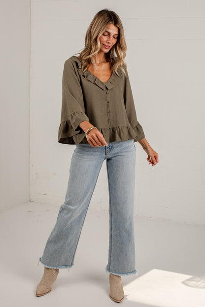 A woman modeling a sage green ruffled blouse with three-quarter bell sleeves and light-wash blue flare jeans, standing in a bright white studio corner.