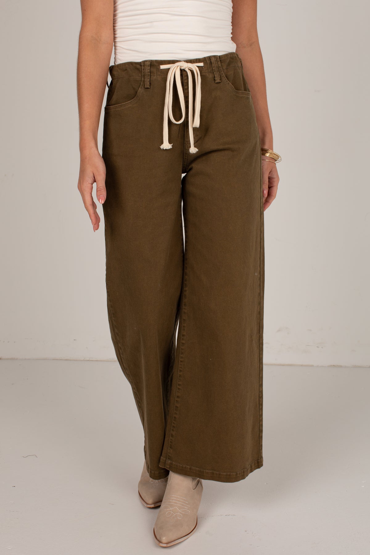 A person wearing wide-leg olive brown pants with a cream drawstring, paired with a white ribbed top and beige ankle boots, shown against a plain light gray studio background.