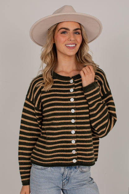 A woman wearing a brown and black horizontally striped knit cardigan sweater with white buttons down the front, styled with a beige wide-brim hat and light-wash blue jeans against a solid gray studio background.