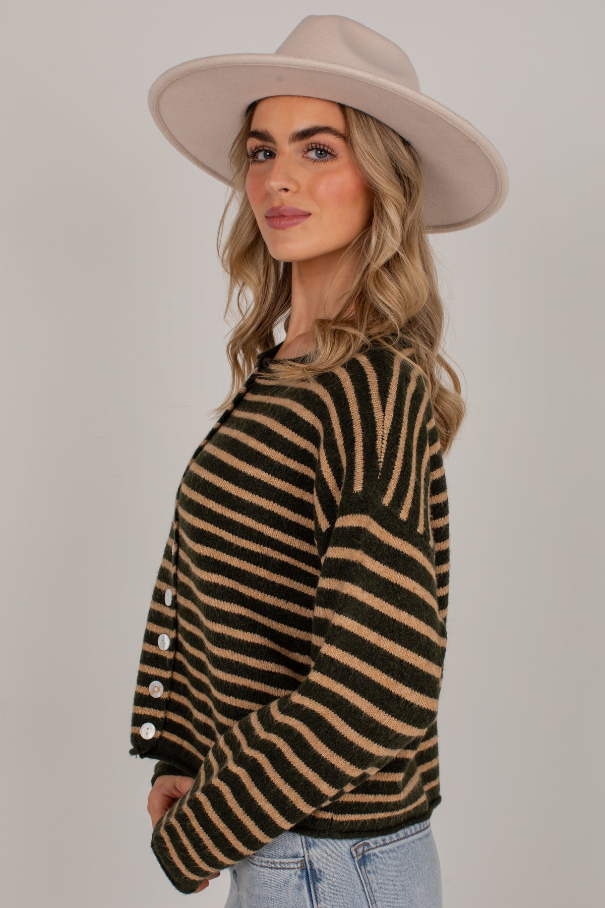A woman wearing a beige wide-brim hat and a brown and beige horizontally striped knit cardigan with white buttons on the sleeve, paired with light blue jeans, photographed against a solid light gray background.