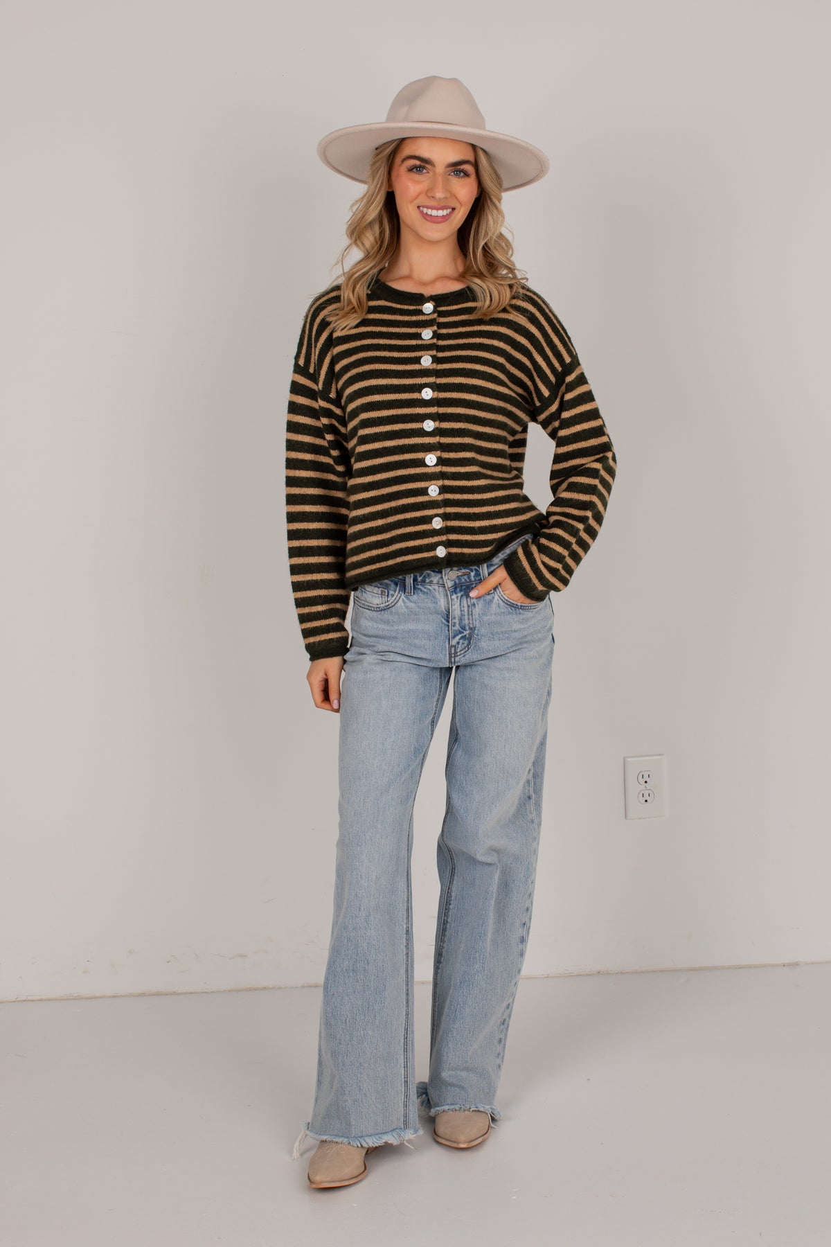 A woman modeling a brown and black striped button-front knit sweater, paired with light-wash flare jeans, beige flats, and a beige wide-brim hat, standing against a plain white wall.