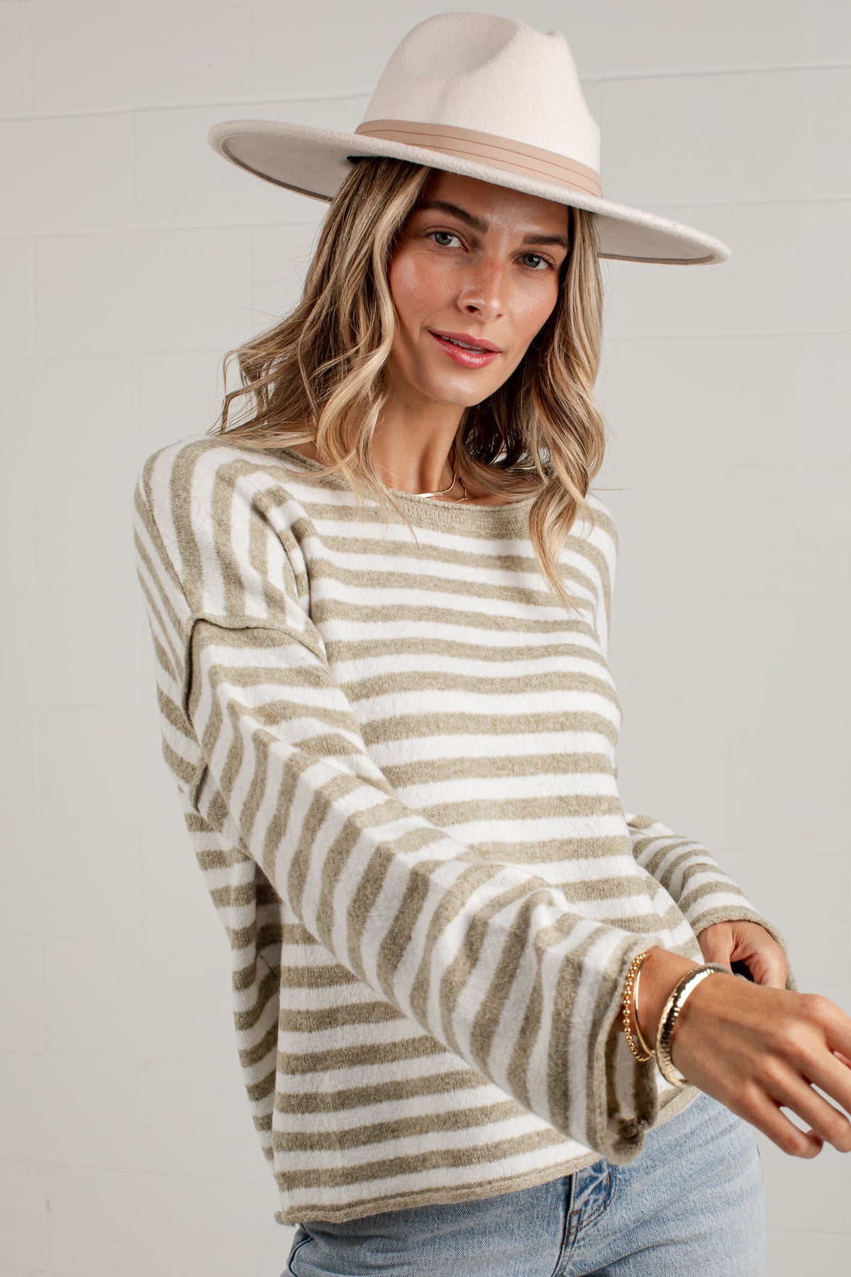 A woman wearing a cream and beige horizontally striped knit sweater, a wide-brimmed cream hat, and light blue jeans, posing against a light gray wall.