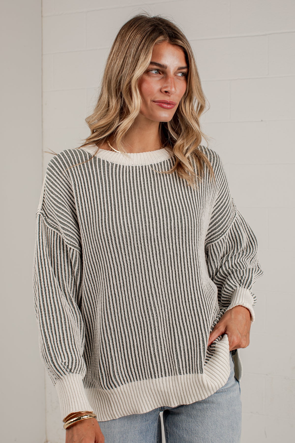 A woman wearing a long-sleeve knit sweater with vertical gray and off-white stripes, paired with light blue jeans, standing against a white paneled wall.