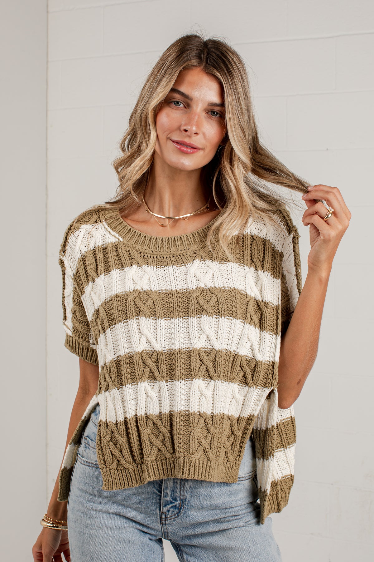 A woman wearing a short-sleeve knit sweater with horizontal beige and off-white stripes, paired with light-wash jeans, standing against a white paneled wall.