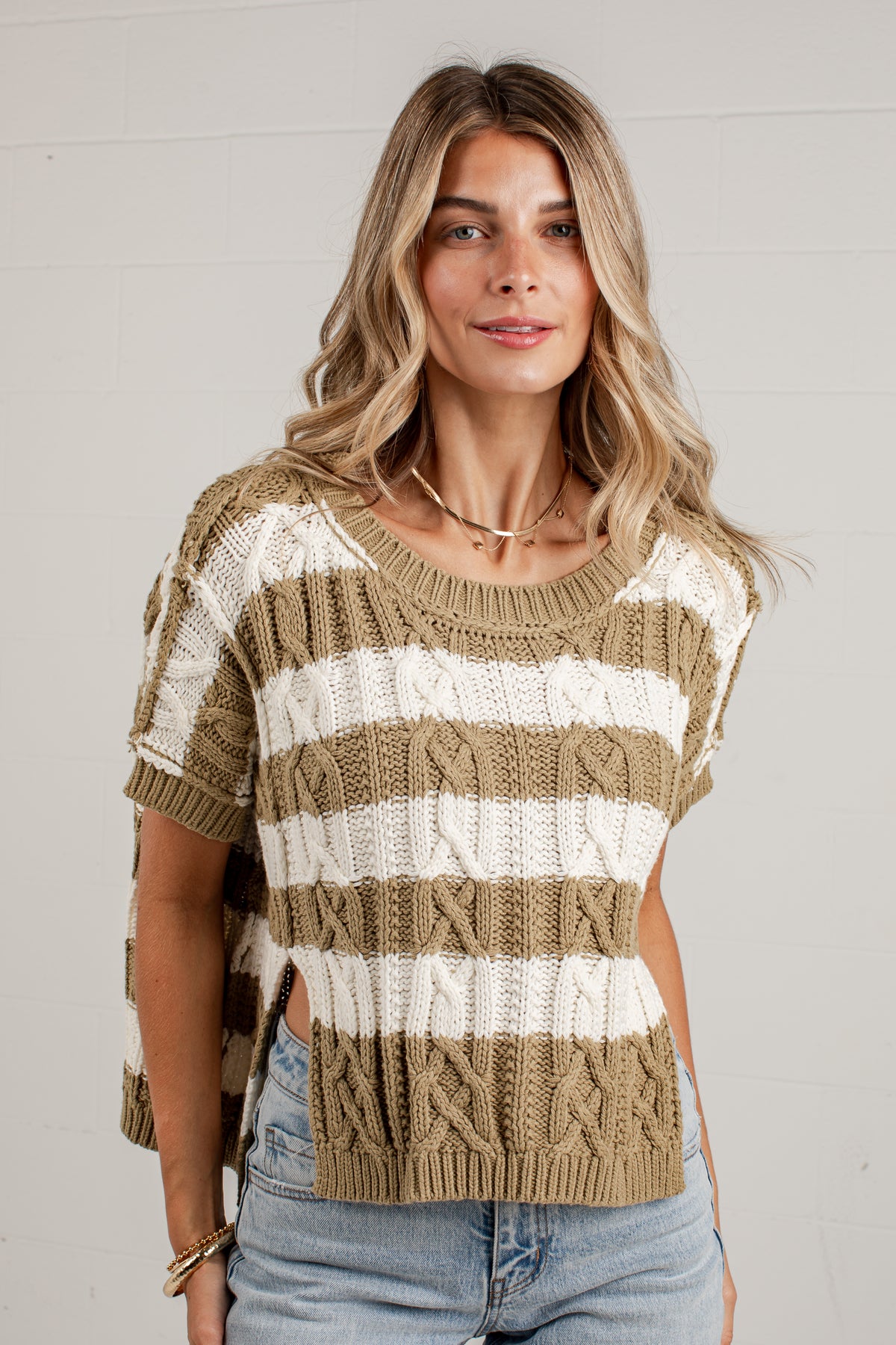A woman wearing a short-sleeve knit sweater with horizontal beige and off-white stripes, paired with light-wash jeans, standing against a white paneled wall.