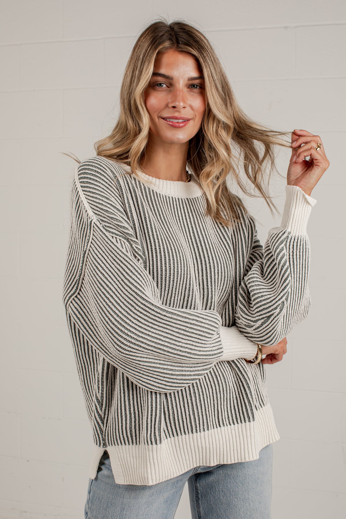 A woman wearing a cream and gray vertically striped knit sweater with balloon sleeves and ribbed cuffs, hem, and collar, paired with light blue jeans against a white paneled wall.