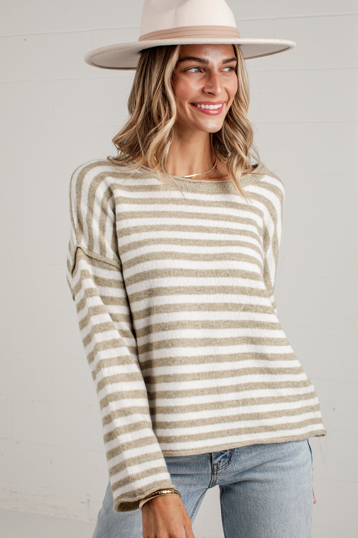 A woman wearing a beige and off-white horizontally striped knit sweater, a wide-brimmed beige hat, and light blue jeans, standing against a light gray wall.