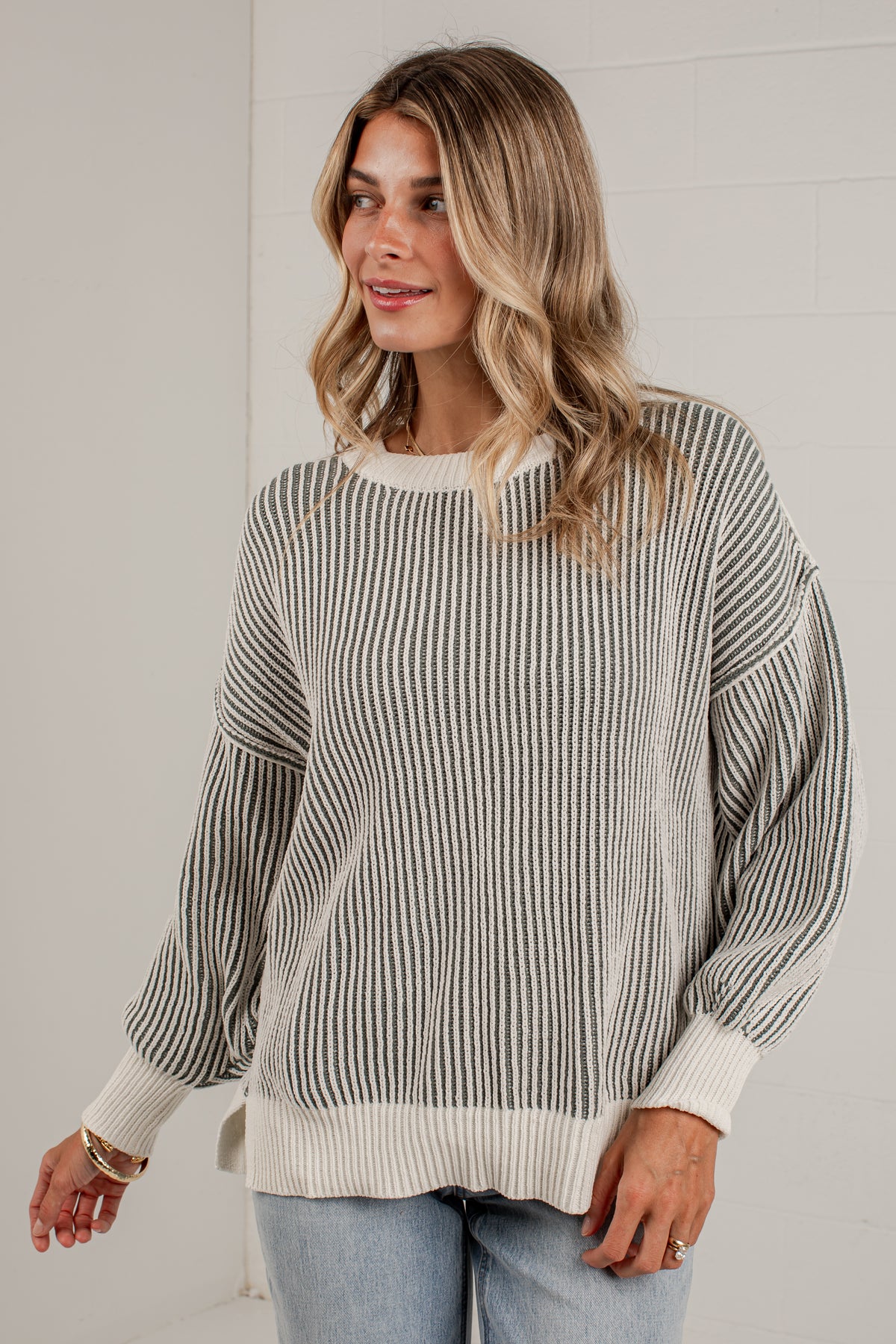 A woman wearing a cream and olive vertically striped knit sweater with balloon sleeves, paired with light blue jeans, standing against a white brick wall.